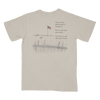 A light beige crewneck t-shirt shown from the back, featuring a black line art graphic of a rural scene with an American flag, fence, grass, and a dog, accompanied by a humorous political text print.