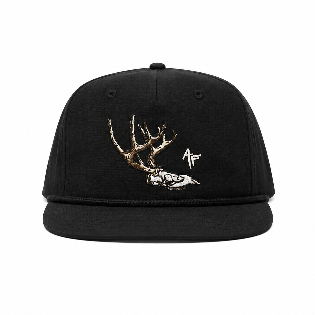 A black flat-brim snapback cap featuring a graphic of a deer skull with antlers and a small white monogram logo on the front panel, set against a plain white background.
