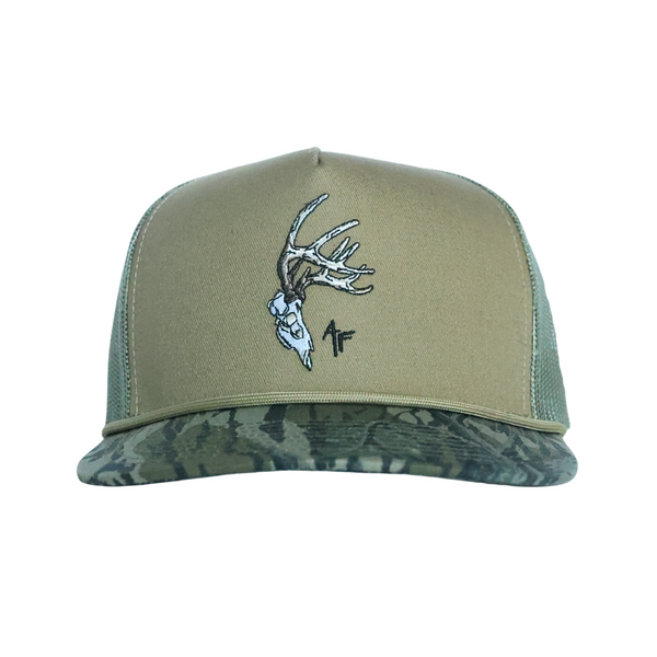 A trucker-style baseball cap with an olive green front panel, matching green mesh back, and a camouflage patterned visor. The front panel features an embroidered deer skull with antlers and a small black  AF' logo.
