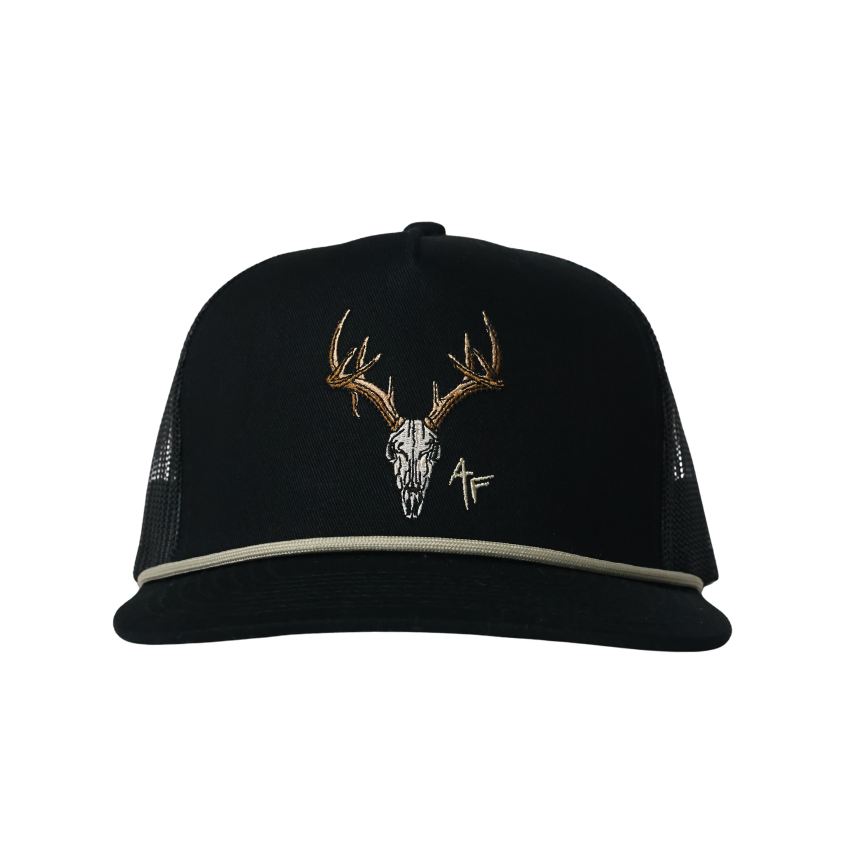 A black trucker hat with a curved brim, featuring an embroidered deer skull with antlers in white, gold, and brown on the front panel, and a thin light gray rope detail along the edge of the brim.
