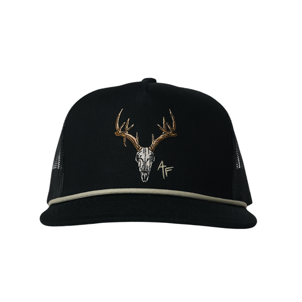 A black trucker hat with a curved brim, featuring an embroidered deer skull with antlers in white, gold, and brown on the front panel, and a thin light gray rope detail along the edge of the brim.