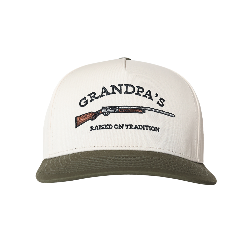 An off-white and olive green trucker-style cap featuring a front graphic with the text 'GRANDPA'S RAISED ON TRADITION' and a detailed illustration of a shotgun.