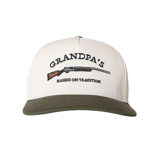 An off-white and olive green trucker-style cap featuring a front graphic with the text 'GRANDPA'S RAISED ON TRADITION' and a detailed illustration of a shotgun.