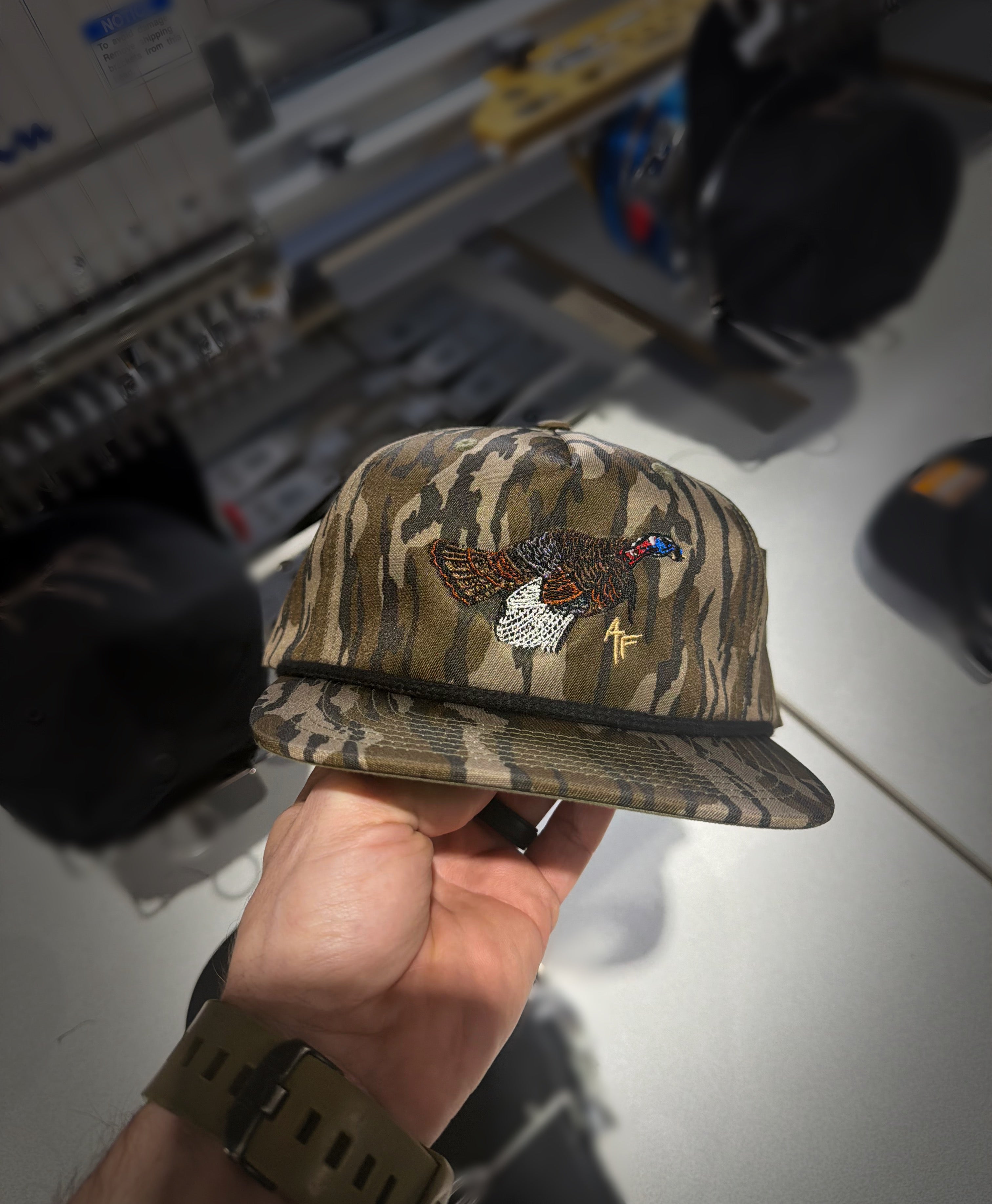 Camouflage cap with embroidered designs held in a hand against a blurred background